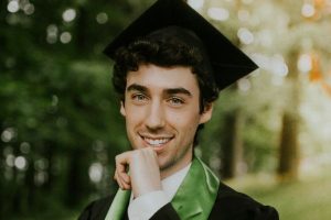 Confident male PhD graduate in cap and gown with a green stole, smiling outdoors in a wooded area, symbolising achievement and new beginnings.