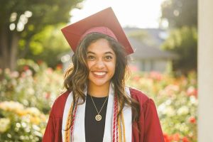 Smiling PhD graduate in a maroon cap and gown standing in a sunlit garden, representing success and confidence after completing a doctoral journey.