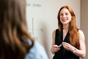 Smiling young woman discussing feedback at a whiteboard, representing confidence and clarity during PhD work.